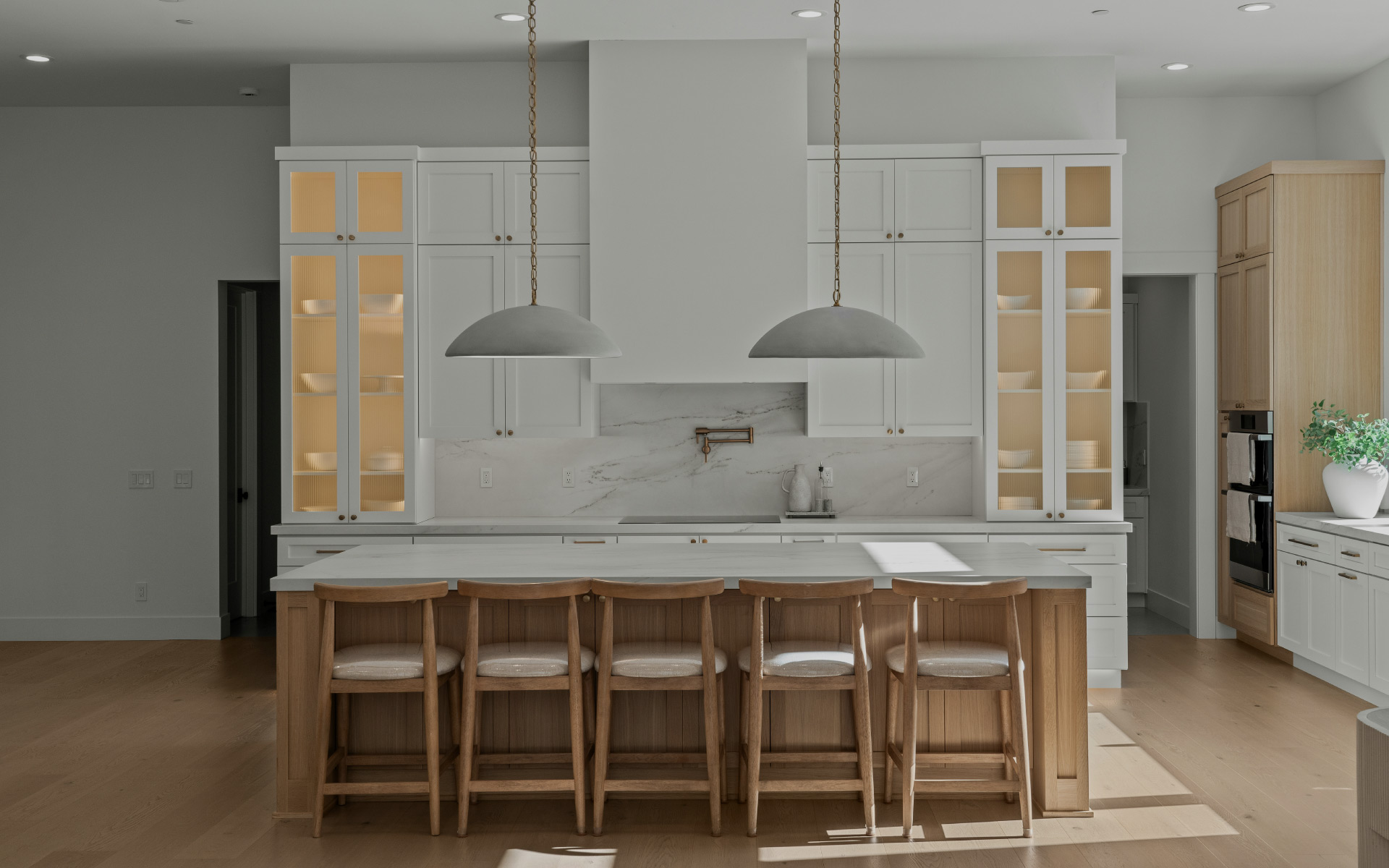 A spacious kitchen featuring a wooden island with five barstools, dome pendant lights overhead, white main cabinetry with lit glass-display units, a white range hood, and light hardwood flooring.