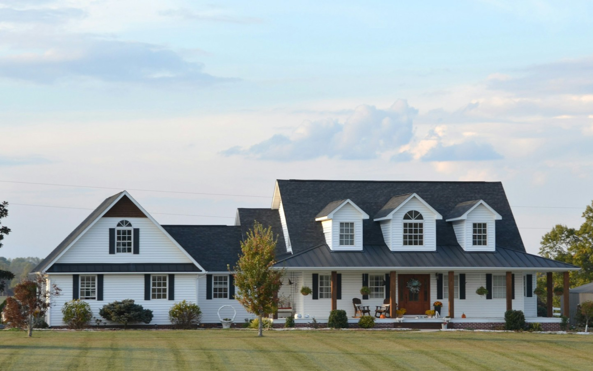 A two-story white farmhouse with black shutters, three dormer windows, a spacious front porch with wooden columns, and a large manicured green lawn.